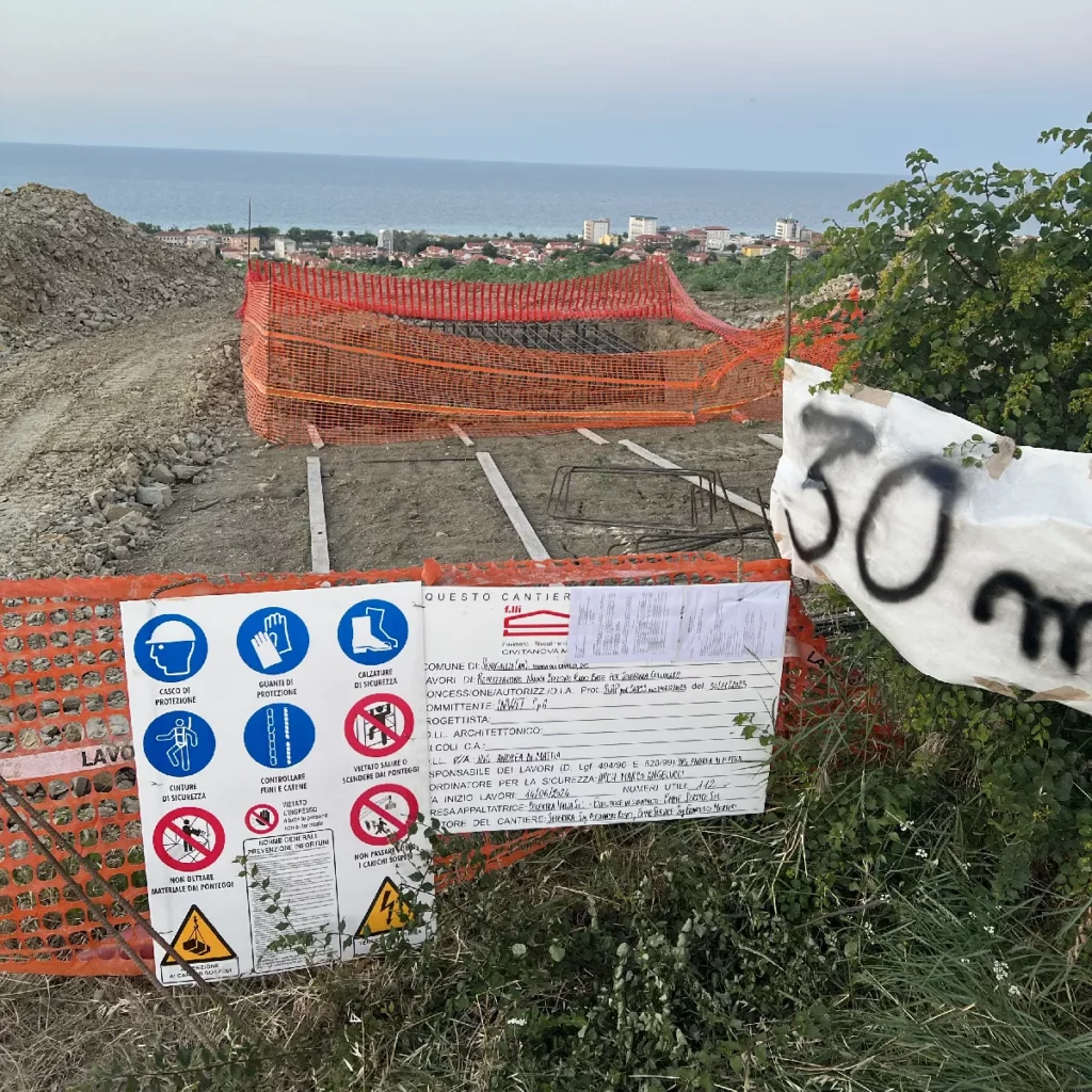 Cartelloni con le autorizzazioni e gli avvisi di pericolo per lavori in corso. Sullo sfondo, le recizioni il buco, la collina e il mare